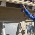 Painter sanding a wood fascia board with an orbital sander, white spot primer already applied to the areas sanded earlier, window below masked in plastic