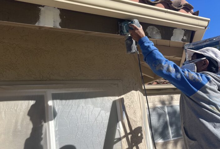 Painter sanding a wood fascia board with an orbital sander, white spot primer already applied to the areas sanded earlier, window below masked in plastic